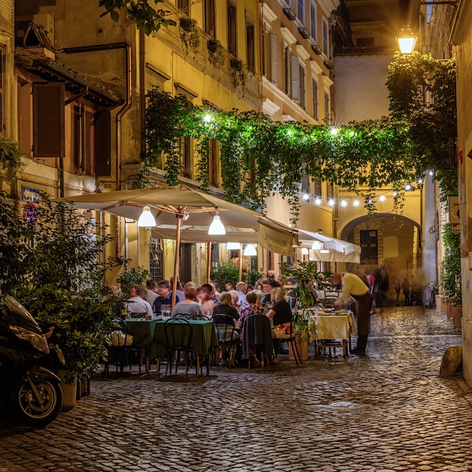 Tables en terrasse d’un restaurant, remplies de clients, dans une rue peu fréquentée de Rome