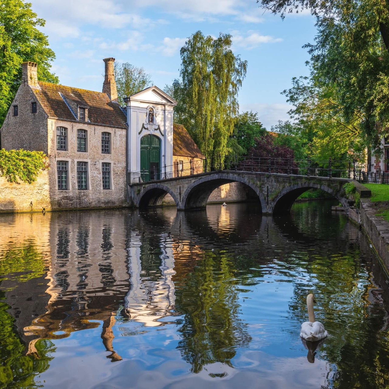 Een sierlijk gebouw staat aan de oever van een rustige rivier, met een brug die naar het groene gras aan de overkant leidt